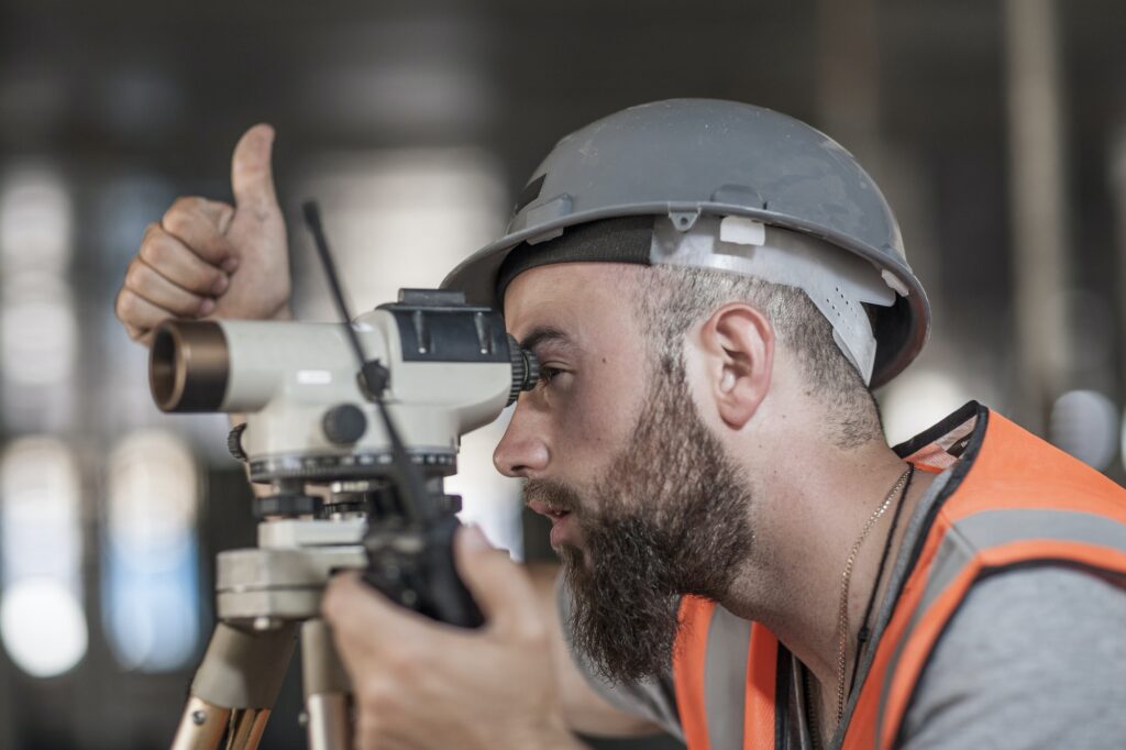 Young male surveyor looking through theodolite giving thumbs up on construction site